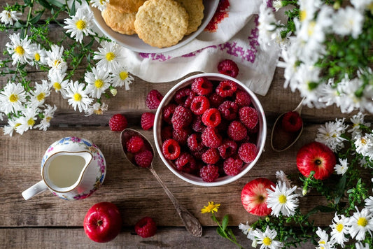 Un bol de framboises fraîches est posé sur une table en bois, entouré de pommes, de biscuits, d'un pot de crème et de fleurs blanches.