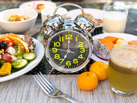 Un réveil analogique argenté se dresse sur une table parmi divers articles de petit-déjeuner, notamment de la salade, des fruits, du jus et un verre de lait.