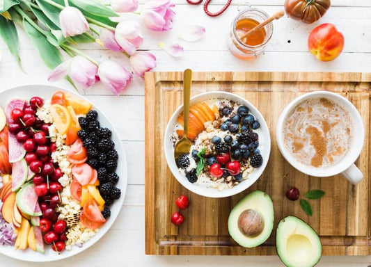 Une scène de petit-déjeuner avec un bol de fruits et de céréales, une assiette avec divers fruits frais, un avocat, une tasse de café, du miel, des tulipes et une pêche sur une surface en bois.