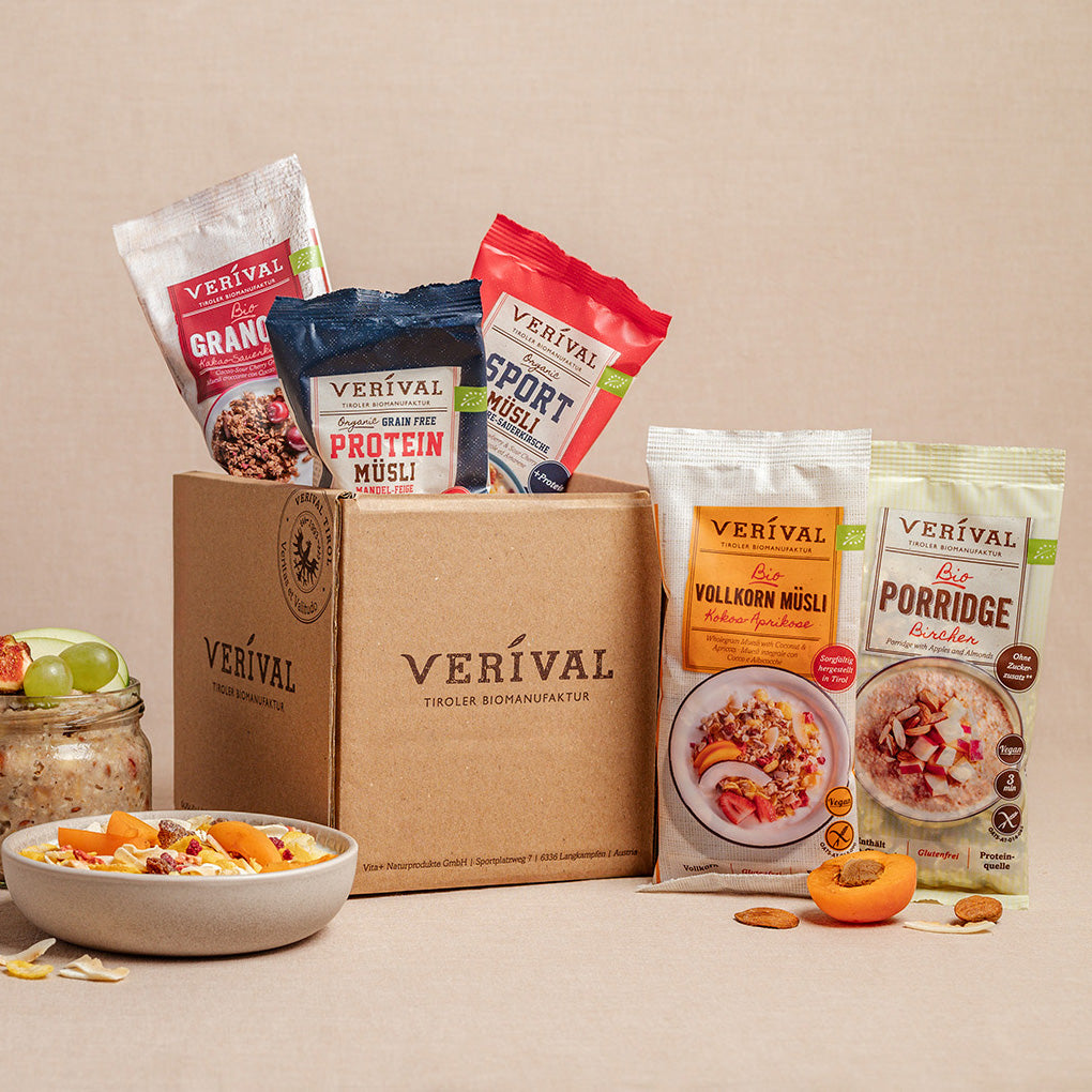 Une boîte avec le logo Verival, entourée de cinq paquets de muesli et de porridge différents, devant lesquels se trouvent deux bols de muesli préparé et de fruits.