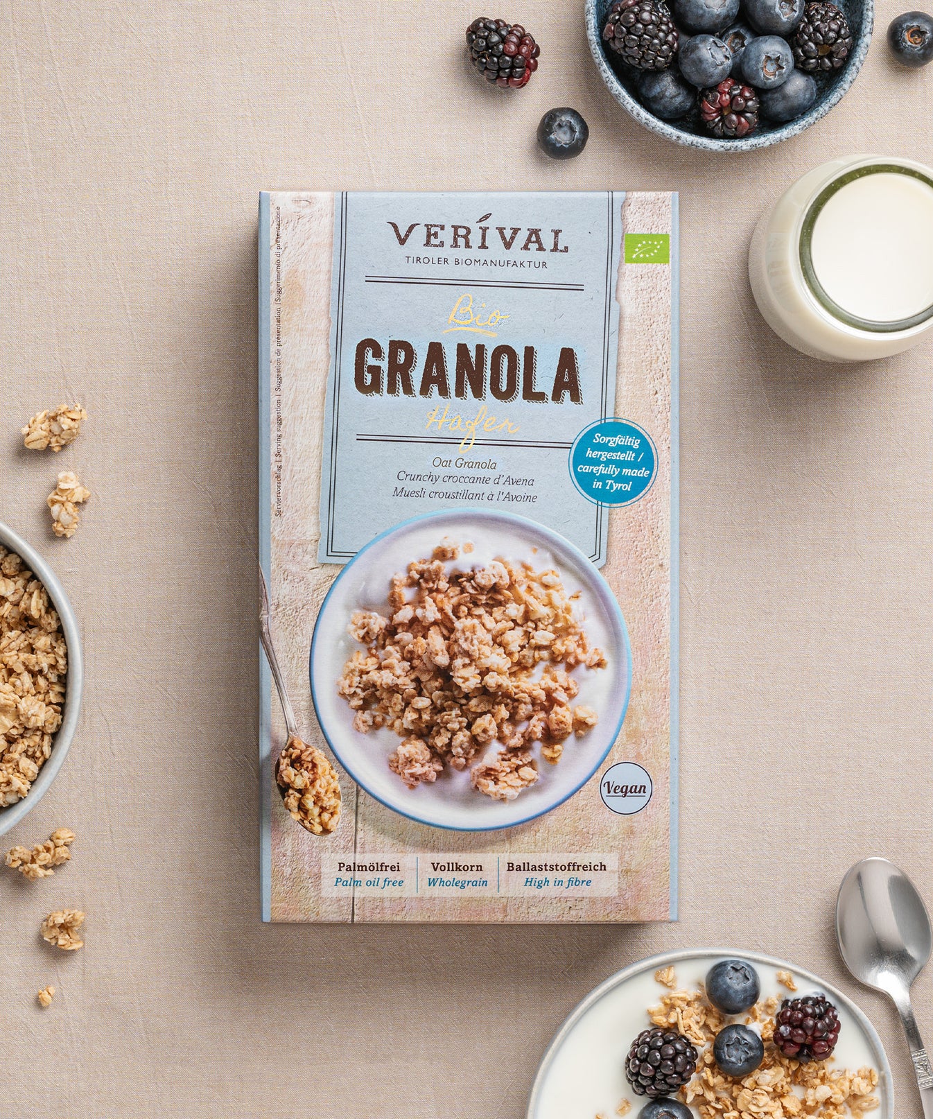 Auf einem beigen Untergrund steht ein Paket Hafer-Granola von Verival Bio, dazu ein Löffel Knusper-Granola, eine Schale Beeren, ein Teller mit Vollkorn-Granola sowie ein Glas Milch.