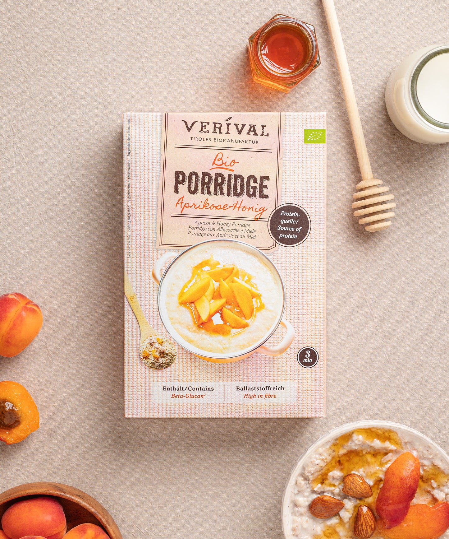 Eine Packung Aprikosen-Honig Porridge von Verival Bio steht mit frischen Aprikosen, Mandeln, einem Honiglöffel, einem Glas Honig und Milch auf dem Tisch.