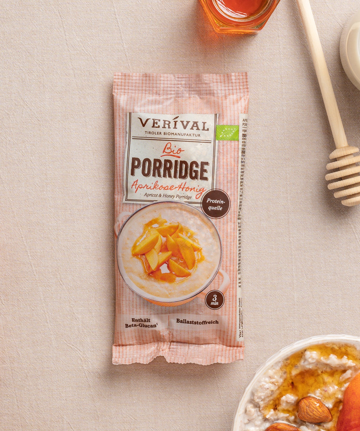 Eine Einzeportinspackung Aprikosen-Honig-Porridge von Verival Bio liegt auf einer beigen Oberfläche neben einer Schüssel cremigen Porridges mit orangefarbenen Fruchtstücken und einem Honiglöffel.