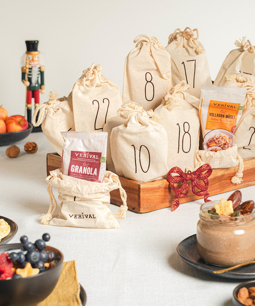 Adventskalender-Säckchen von Verival Bio, nummeriert von 1 bis 24, beinhalten eine reiche Auswahl an Granola-, Porridge- und Müsli-Einzelportionen mit Bio-Zutaten. Ein Nussknacker aus Holz, Obst im Hintergrund und ein Schokoladenmousse-Dessert stehen daneben auf dem Tisch.