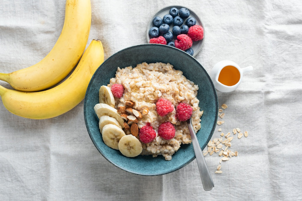 Un bol de porridge avec des tranches de banane, des framboises et des noix, à côté deux bananes, une assiette de myrtilles et de framboises, un petit pot de miel et des flocons d'avoine éparpillés sur un chiffon.