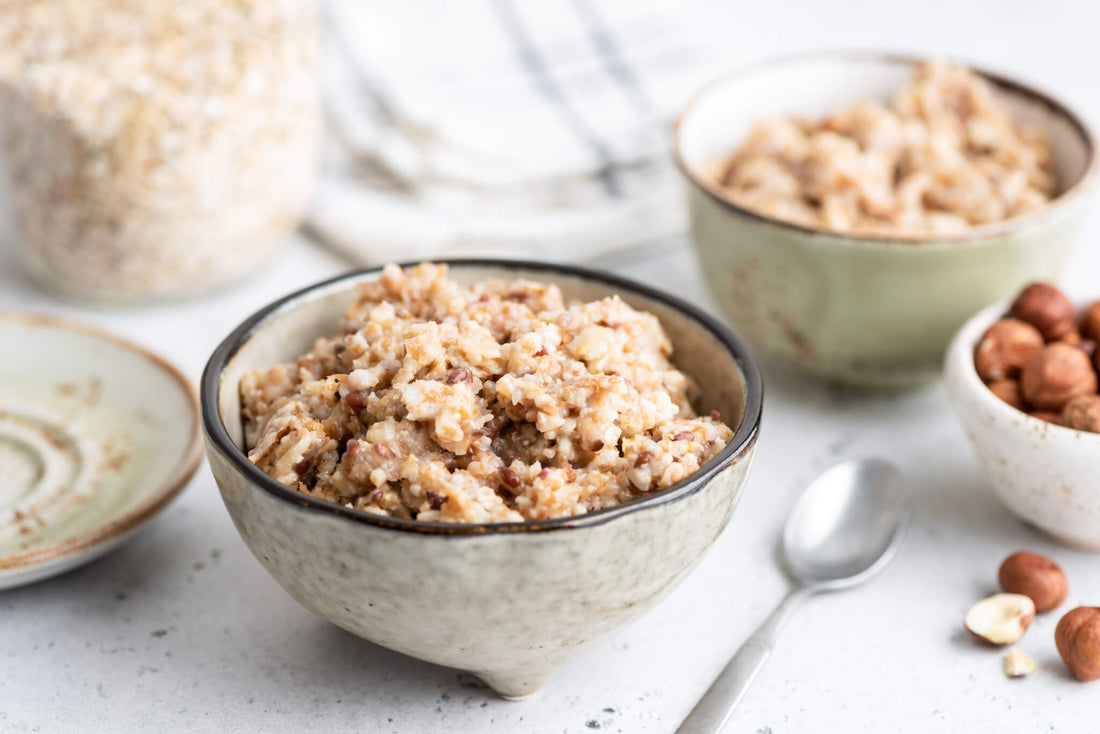Sur une table se trouvent un bol en céramique rempli de porridge à côté d'une cuillère, un autre bol de porridge, une assiette et un petit bol de noisettes.