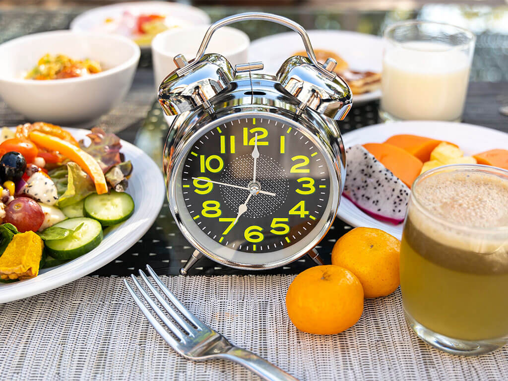 Un réveil analogique argenté se dresse sur une table parmi divers articles de petit-déjeuner, notamment de la salade, des fruits, du jus et un verre de lait.