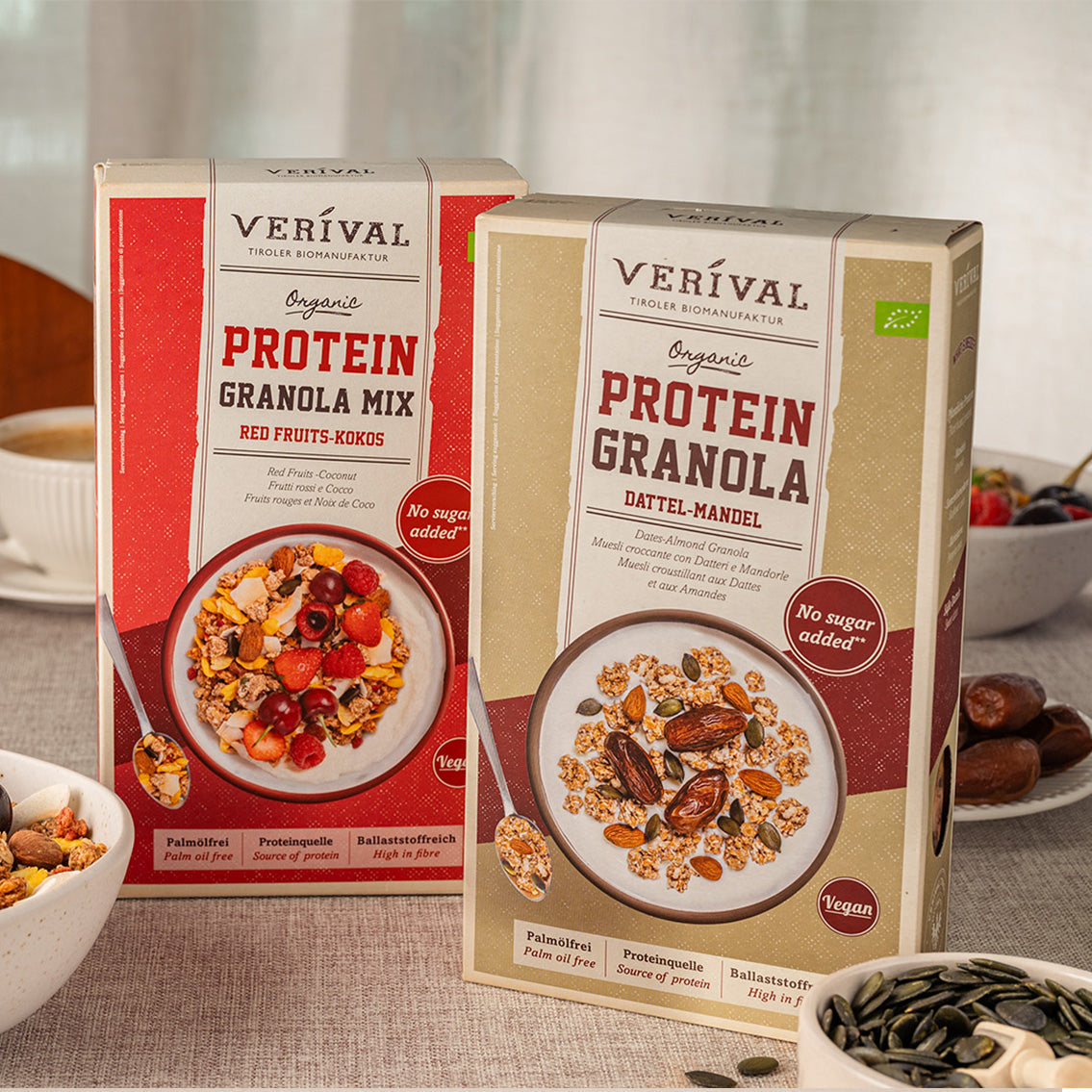 Deux boîtes de granola protéiné biologique Verival, l'une avec des fruits rouges et de la noix de coco et l'autre avec des dattes et des amandes, sont posées sur une table avec des bols de granola et des ingrédients.