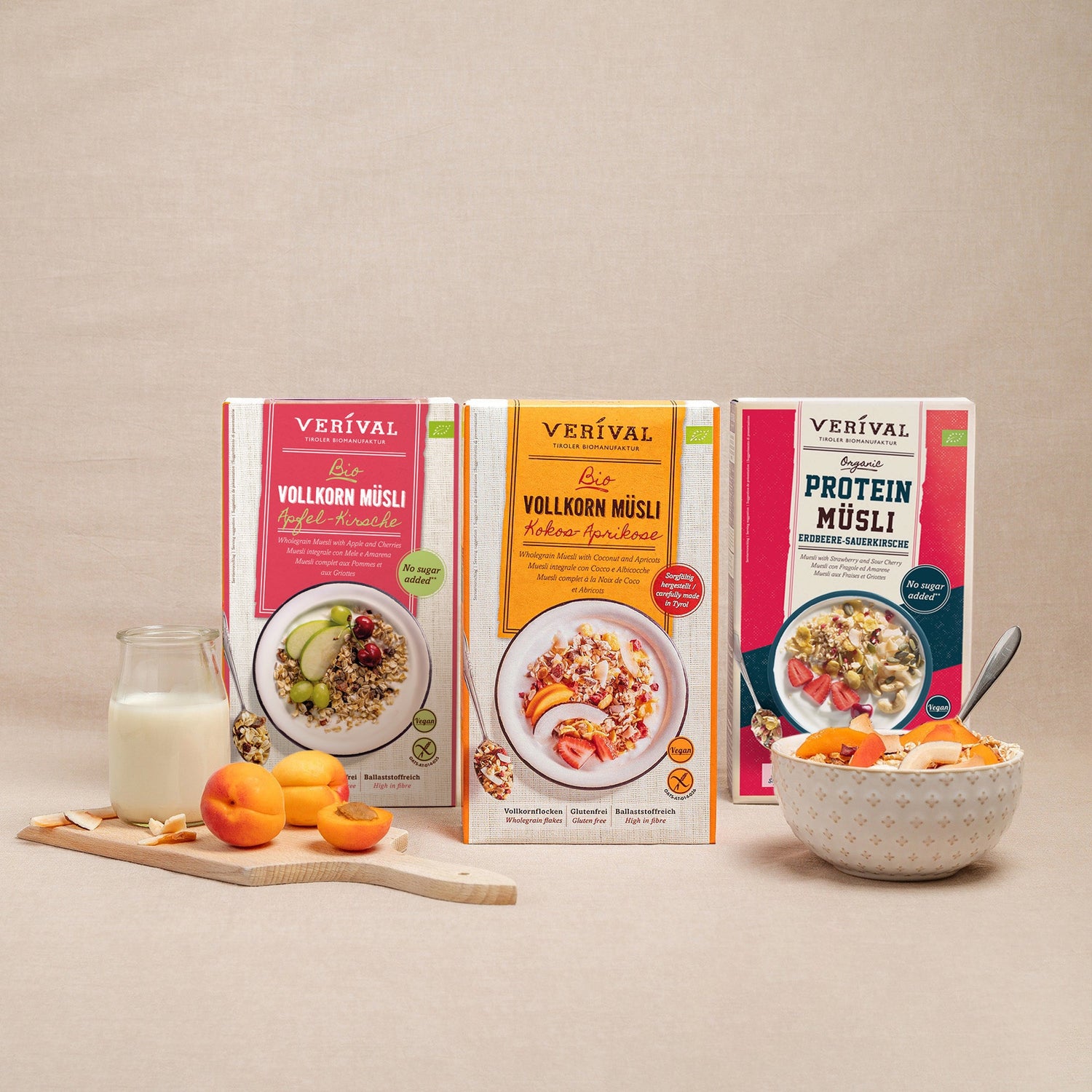 Trois boîtes de muesli complet biologique Verival aux saveurs variées se dressent debout sur une surface beige, à côté d'un bol de muesli, de tranches de pomme, de raisins et d'un verre de lait.