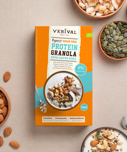 Eine Packung Bio Nuss-Kokos Grain Free Protein Granola von Verival Bio GmbH, umgeben von kleinen Schälchen mit Mandeln, Kürbiskernen und Kokosflocken sowie einem Schälchen Joghurt, das mit dem pflanzlichen Protein-Müsli garniert ist.
