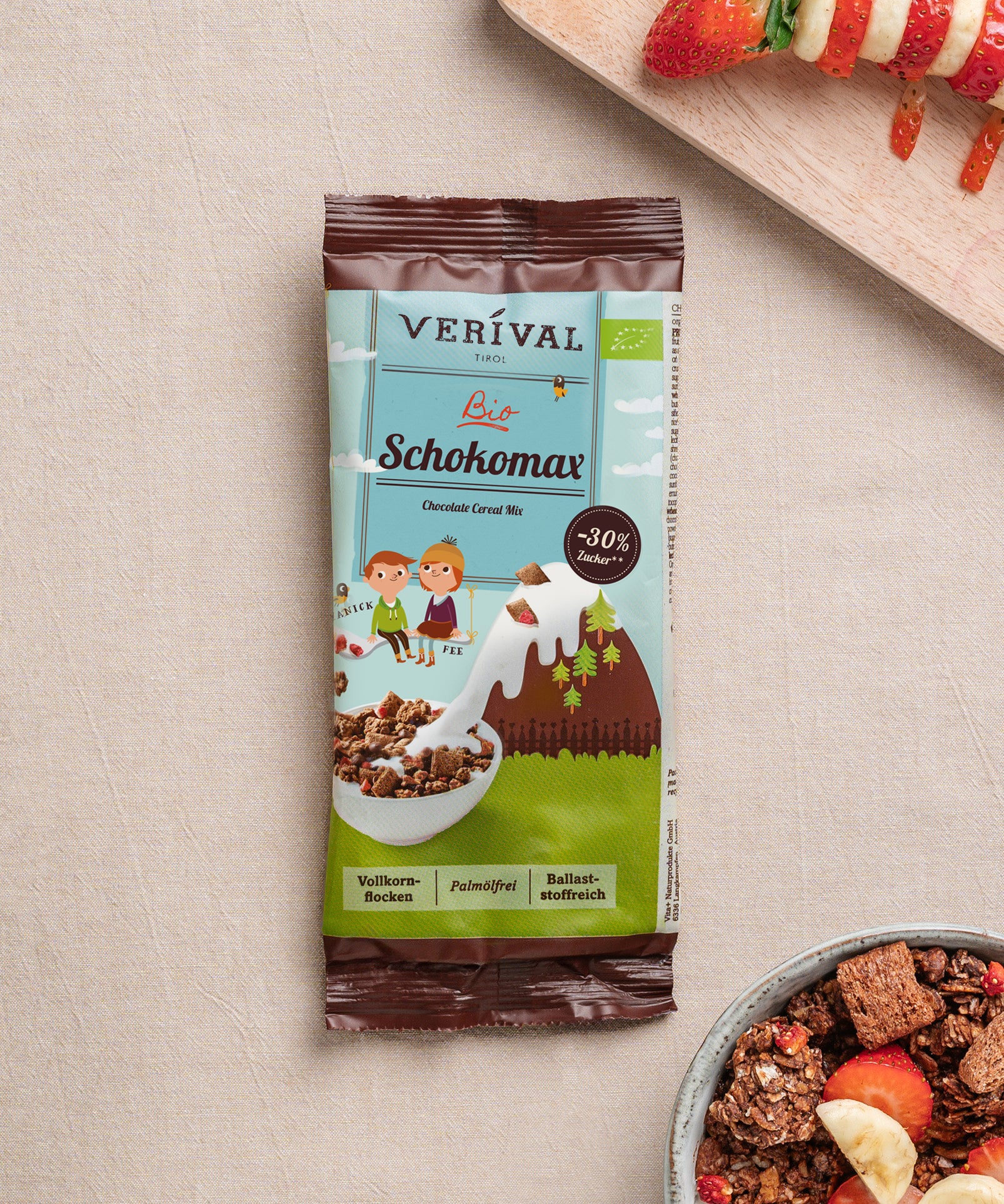 Auf einem beigen Untergrund liegt ein Bio Schokomax Schokoriegel der Verival Bio GmbH neben einer Schale Knuspermüsli mit Früchten und einem teilweise abgebildeten Teller mit Fruchtspießen.