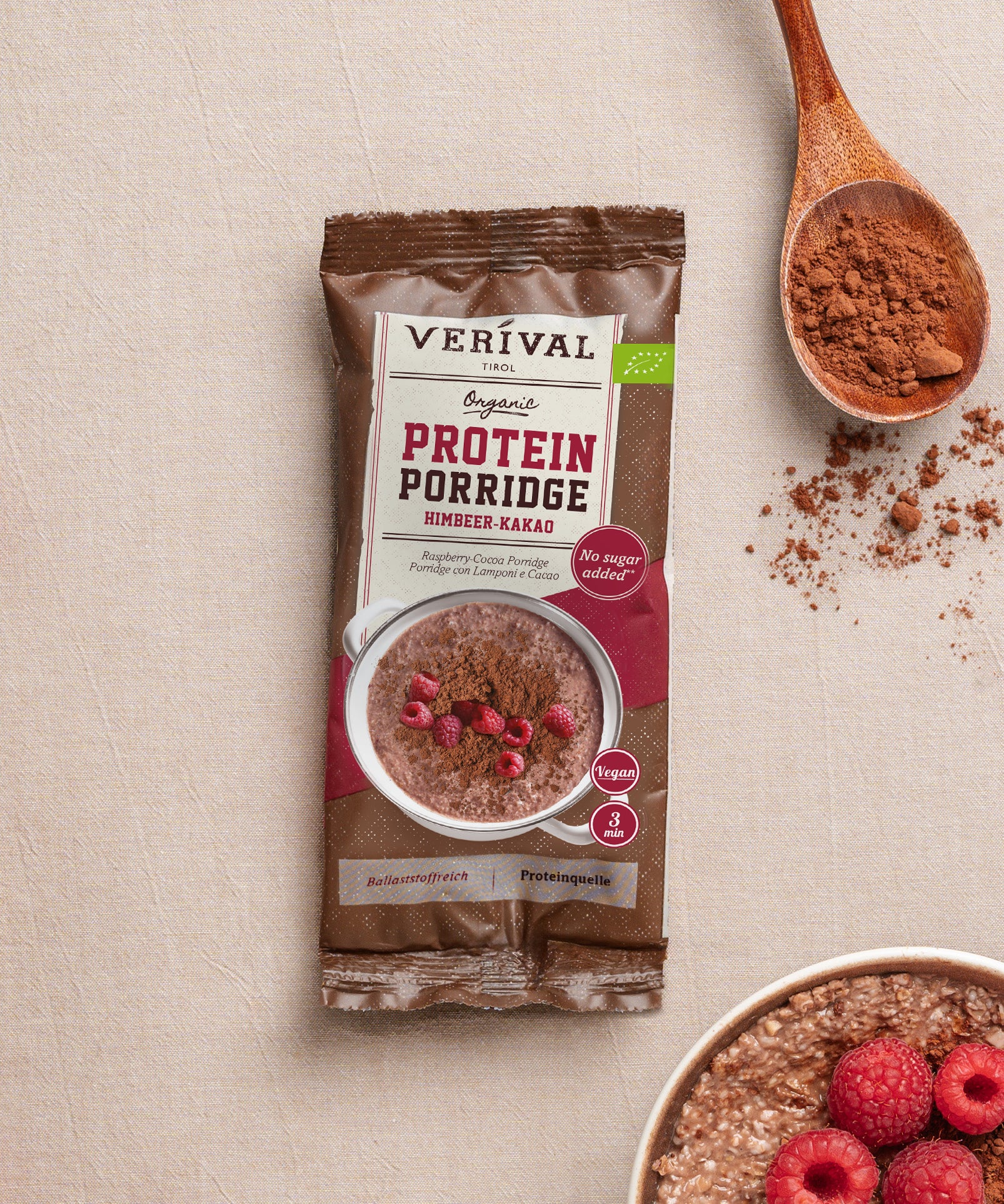 Eine Packung Bio Protein Porridge Himbeer-Kakao der Verival Bio GmbH liegt auf einer beigen Oberfläche neben einem Löffel Kakaopulver, frischen Himbeeren und einer Schüssel ballaststoffreichem veganen Frühstück.