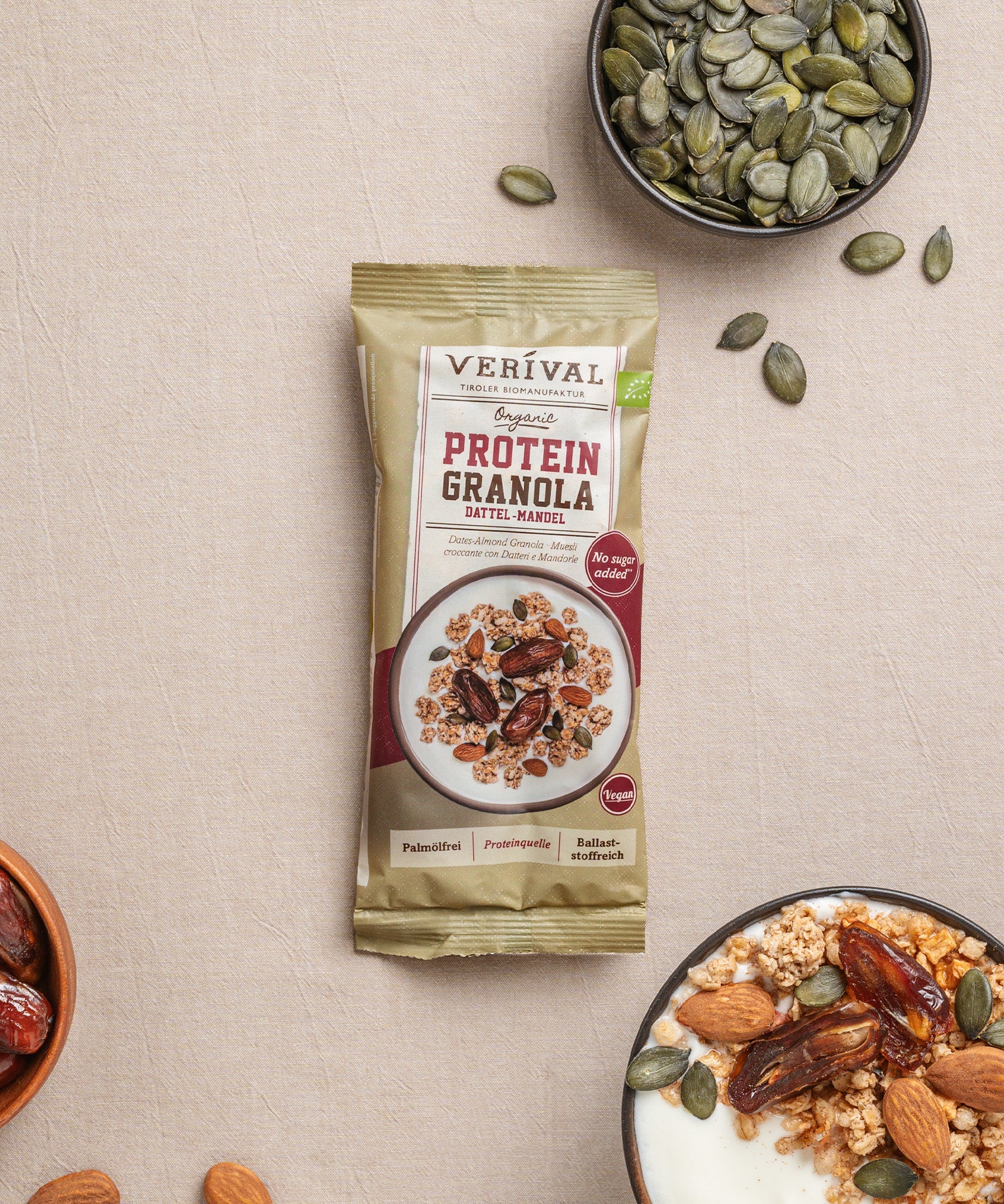 Eine Enzelportionspackung Protein-Granola Dattel-Mandel von Verival Bio steht neben einer Joghurtschale mit Granola, Mandeln, Datteln und Kürbiskernen in einer kleinen Schale.