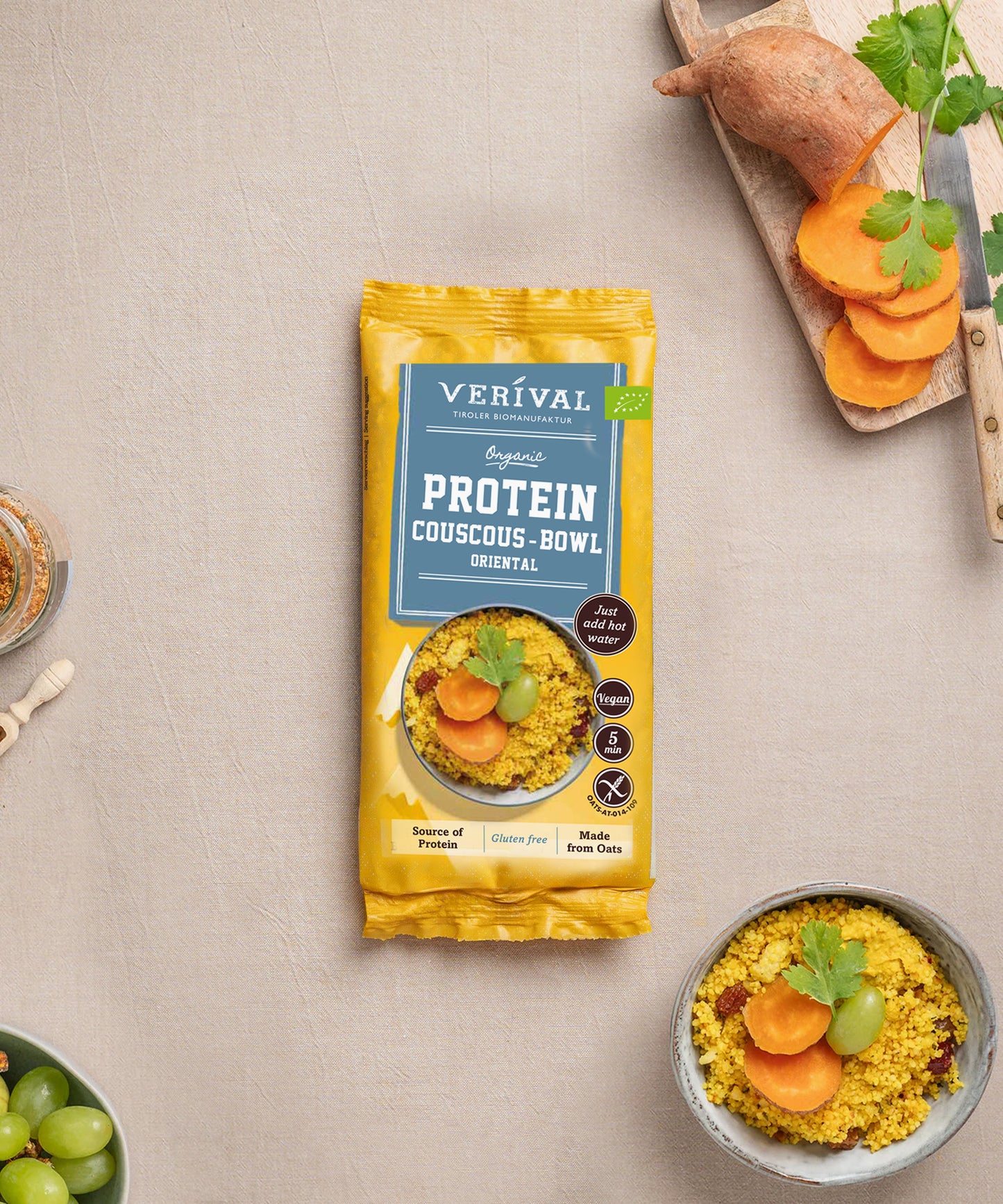 Eine Einzelportionspackung Protein Couscous Bowl Oriental von Verival Bio steht neben einer Schüssel zubereitetem Couscous mit Süßkartoffelscheiben, Weintrauben und Koriander – die perfekte Wahl für ein gesundes Mittagessen voller pflanzlicher Proteine.