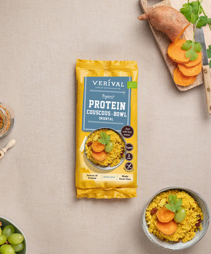 Eine Einzelportionspackung Protein Couscous Bowl Oriental von Verival Bio steht neben einer Schüssel zubereitetem Couscous mit Süßkartoffelscheiben, Weintrauben und Koriander – die perfekte Wahl für ein gesundes Mittagessen voller pflanzlicher Proteine.