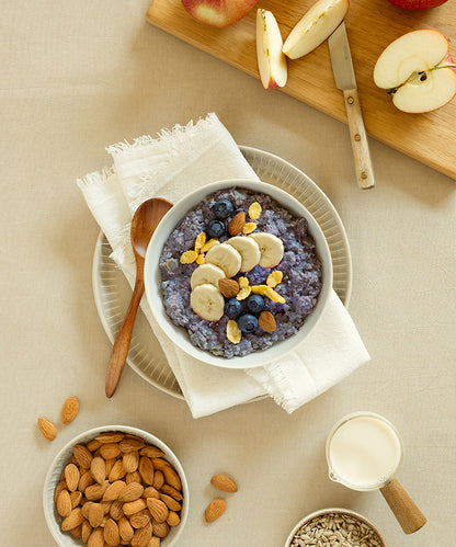 Eine Schüssel „Bio High Protein Porridge Heidelbeere-Banane“ von Verival Bio GmbH mit Banane, Heidelbeeren und Müsli wird mit einem Löffel serviert. Daneben liegen Mandeln, Apfelscheiben auf einem Brett, Sonnenblumenkerne und ein Krug Bio-Sojamilch.