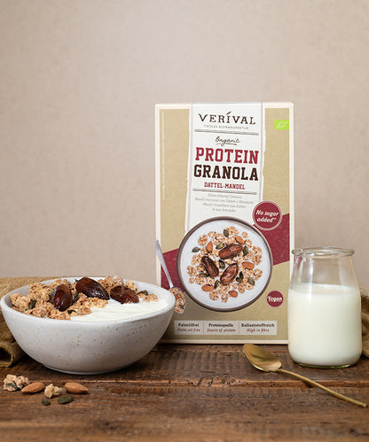 Eine Schüssel Bio-Dattel-Mandel-Protein-Müsli von Verival Bio GmbH, garniert mit Datteln und Mandeln, neben einer Packung des ballaststoffreichen Müslis und einem Glas Milch.